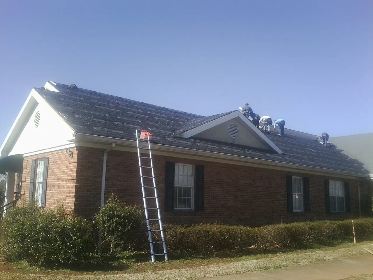 Crew tearing shingles off a brick ranch roof with a ladder set — Metal Roof Repair work by our crew in Auburndale